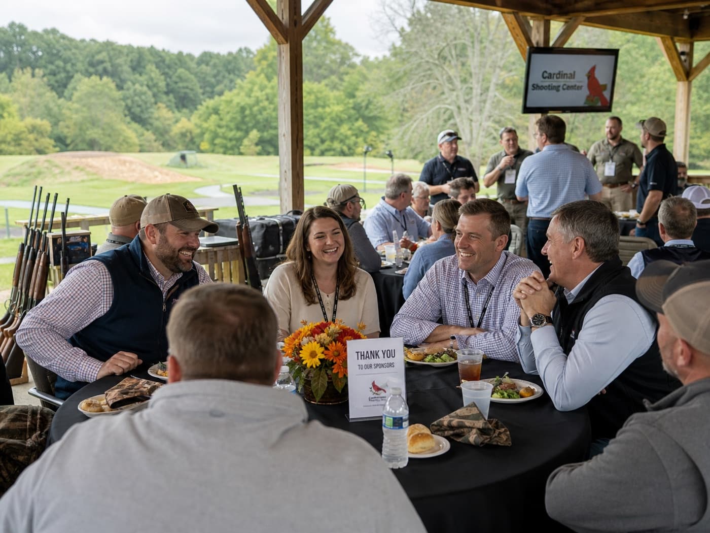 Corporate sponsor lunch under the pavilion at The Cardinal Center, with a 'Thank You to Our Sponsors' table card and shotguns racked in the background.